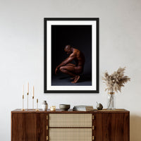 Framed artwork hanging above a brown walnut credenza, the art is a fine art photo showing a sideview of a nude black man crouching with arms resting on knees and head tilted down.