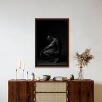 Framed canvas artwork hanging above a brown walnut credenza, the art is a black and white fine art photo showing a sideview of a nude black man crouching with arms resting on knees and head tilted down.