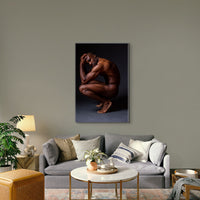 Photo on canvas artwork hanging above the sofa in a boho-glam style livingroom, the art is showing showing a sideview of a nude black man crouching with elbows on knees, hands on head, looking at the camera.