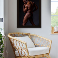 A rattan chair in a bright corner of a bedroom with artwork hanging on the wall above the chair, the art is a framed canvas photo of the sideview of a nude black man from head to mid thigh with one arm bent and chin resting on closed fist.