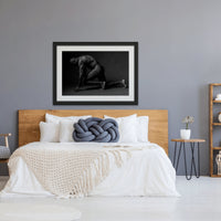 Framed artwork hanging above a light coloured wood headboard in a bedroom with natural and blue tones, the art is a black and white photograph of a sideview of a nude black man from head to mid thigh with one arm bent and chin resting on closed fist.