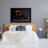Framed canvas artwork hanging above a light coloured wood headboard in a bedroom with natural and blue tones, the art is a photograph of a sideview of a nude black man from head to mid thigh with one arm bent and chin resting on closed fist.