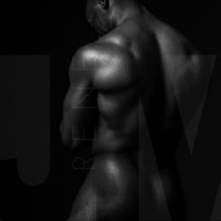 Black and white fine art photo of nude black man posterior view from head to mid thigh with arms crossed, looking downwards.