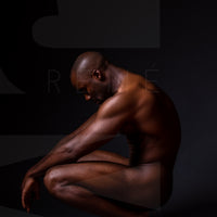 Fine art photo showing a sideview of a nude black man crouching with arms resting on knees and head tilted down.