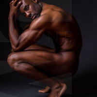 Fine art photo showing a sideview of a nude black man crouching with elbows on knees, hands on head, looking at the camera.