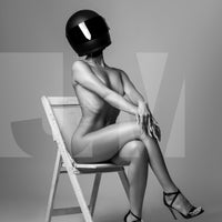 Black and white photo of nude woman wearing a black motorcyle helmet and black high heels sitting on chair with legs crossed, back arched and arms straight with hands resting on knee.