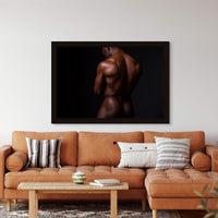 Fine art photo hanging above a brown leather sofa in a modern livingroom of a nude black man posterior view from head to mid thigh with arms crossed, looking downwards.