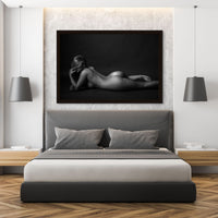 A clean and modern bedroom featuring a framed nude portrait of a woman lying on her stomach with her elbow bent and hand close to her face with is partially obstructed by her long hair. The model has a bent leg and has slightly rolled her hips to highlight the curve of her lower back, hips and buttocks.