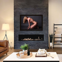 Framed fine art photo hanging above a fireplace in a stylish casual livingroom showing the sideview of a naked woman kneeling and leaning forward with her fingers tented in front of her knees. The woman has long hair which is partially covering her face. The light is shining on her shoulder as well as creating a highlight that follows the curve of her hip and thigh.