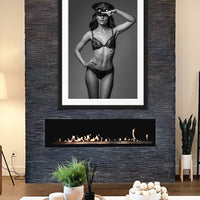 Framed fine art photo hanging above a fireplace in a stylish casual livingroom showing a black and white image of a gorgeous woman wearing a lace bra and panties with a steampunk style captain's hat and round goggles on her head with one hand on hip and the other saluting the camera.