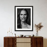 Framed artwork hanging above a brown walnut credenza, the art is a black and white close up portrait of a beautiful woman holding a bullet between her teeth with her brow furrowed.