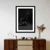 Framed artwork hanging above a brown walnut credenza, the art is a black and white photo showing a sideview of a naked woman in a shoulder stand with arms bent covering her face and knees bent with legs crossed.