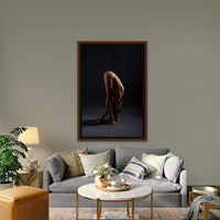 Framed artwork hanging above a brown walnut credenza, the art is a black and white photo showing a sideview of a naked woman in a shoulder stand with arms bent covering her face and knees bent with legs crossed.