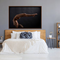 Framed canvas artwork hanging above a light coloured wood headboard in a bedroom with natural and blue tones, the art is a photograph of a sideview of a nude woman in a handstand pose with back arched in a sweeping curve with legs stretching out behind her.
