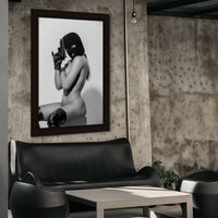 Framed canvas artwork hanging above a black leather couch in an industrial style living room, the artwork is a photo of a seated nude woman, the woman is wearing black thigh high boots, a black balaclava, black leather gloves and is holding a handgun.