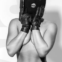 Black and white photo of a nude woman wearing a black balaclava and black gloves holding a handgun, with her hands on her face and the gun resting on the side of her face.