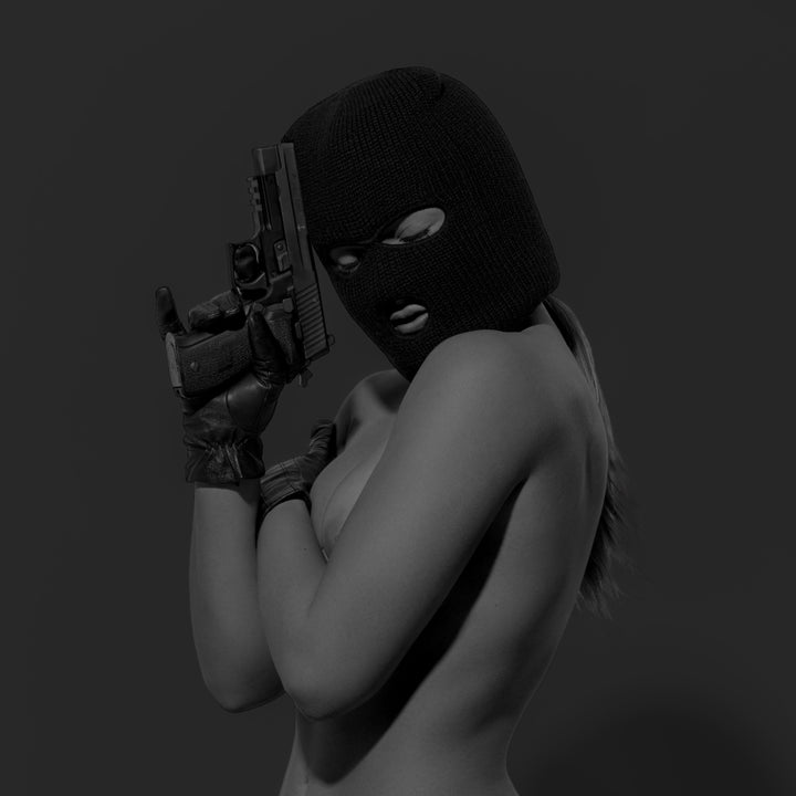 Black and white image of a nude woman from the waist up, the woman is wearing a black balaclava, black leather gloves and is holding a handgun.