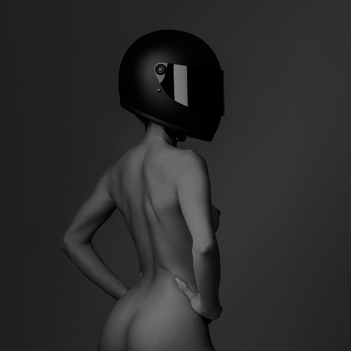 Black and white photo of confident nude woman wearing a black motorcyle helmet standing with back to camera looking over shoulder.