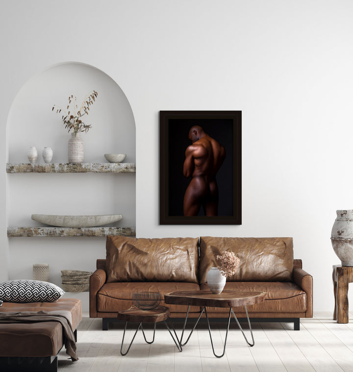 Fine art photo hanging above a brown leather sofa in a modern livingroom of a nude black man posterior view from head to mid thigh with arms crossed, looking downwards.