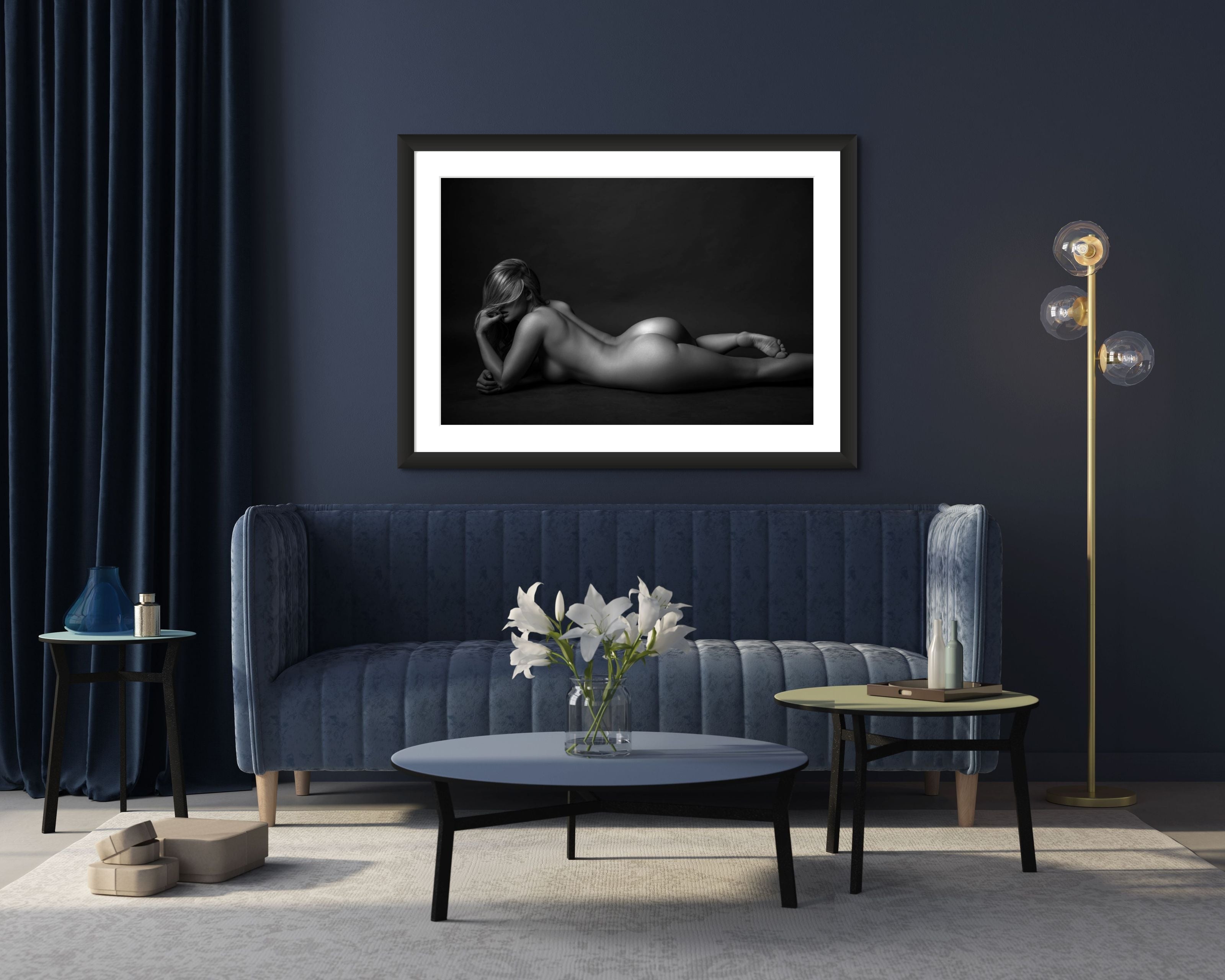 Photo of a living room decorated in deep rich blues featuring a framed piece of art hanging above the sofa. The artwork is a black and white photo of a nude lying on her stomach with one arm bent and her head resting on her chin looking towards the camera, but her hair is swept across her face. The light and shadows accent the curves of her body. 