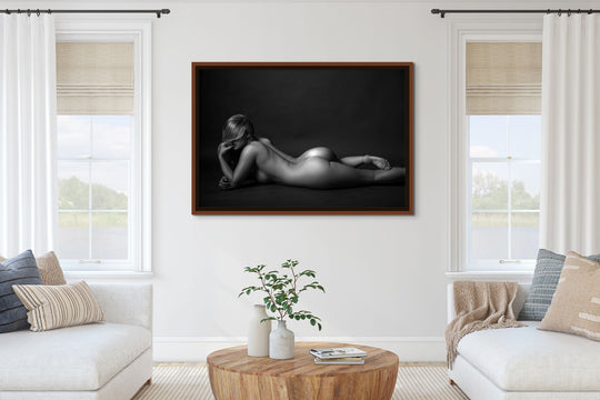 Black and white framed portrait of a nude woman hanging between two windows in a bright neutral living room.