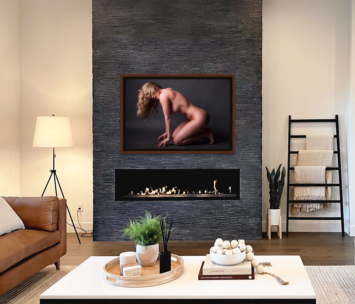 Framed fine art photo hanging above a fireplace in a stylish casual livingroom showing the sideview of a naked woman kneeling and leaning forward with her fingers tented in front of her knees. The woman has long hair which is partially covering her face. The light is shining on her shoulder as well as creating a highlight that follows the curve of her hip and thigh. 