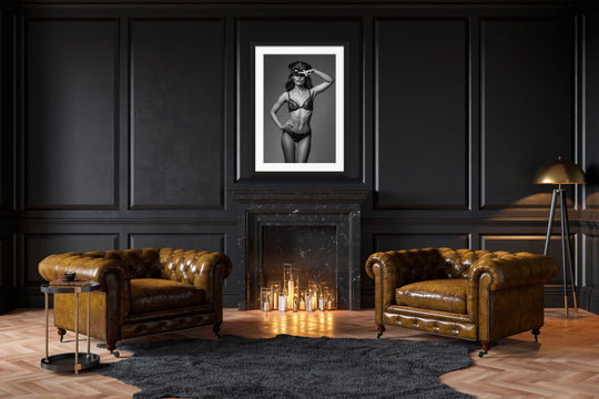 Dark and moody study with fireplace and brown leather chairs. Hanging above the fireplace is a framed portrait of a woman in lace underwear, wearing a steampunk style hat and goggles on her head with her hand on hip and saluting the camera.