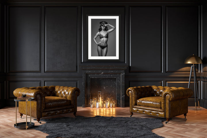 Dark and moody study with fireplace and brown leather chairs. Hanging above the fireplace is a framed portrait of a woman in lace underwear, wearing a steampunk style hat and goggles on her head with her hand on hip and saluting the camera.