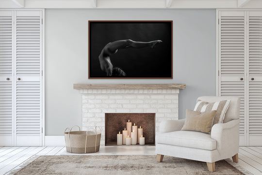 Photo of a living area in a house with a fireplace and chair. Hanging above the fireplace is a framed portrait of a nude woman taken in studio posing in a handstand with her back arched so her lower half is parallel to the ground.