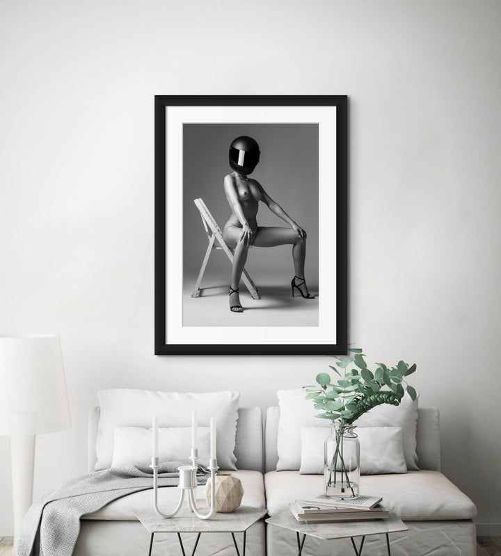 Photo of a bright neutral livingroom featuring framed artwork hanging above the sofa. The artwork is a portrait of a nude woman wearing only black high heels and a blacked out motorcycle helmet. The woman is sitting on a wooden chair in a strong and confident pose.