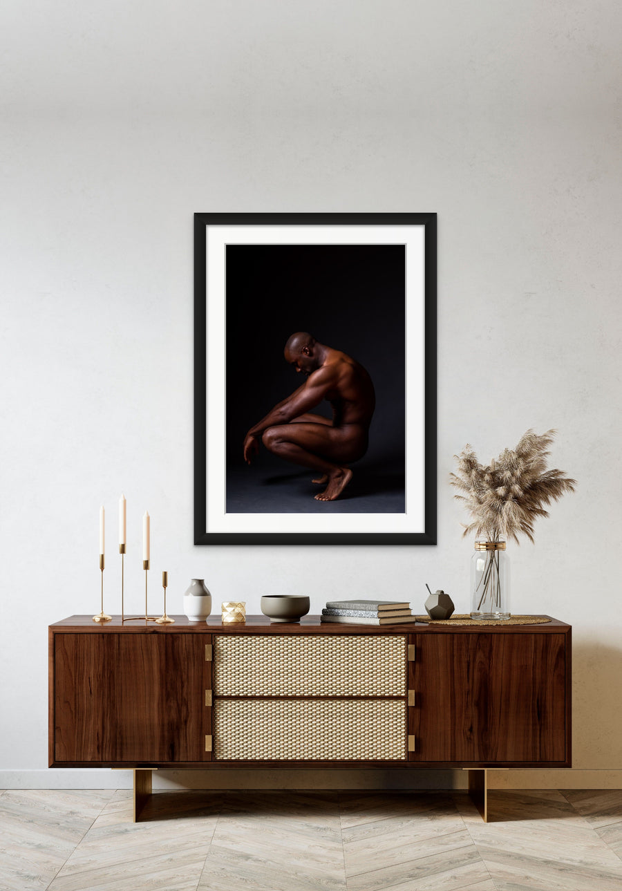 Framed artwork hanging above a brown walnut credenza, the art is a fine art photo showing a sideview of a nude black man crouching with arms resting on knees and head tilted down.