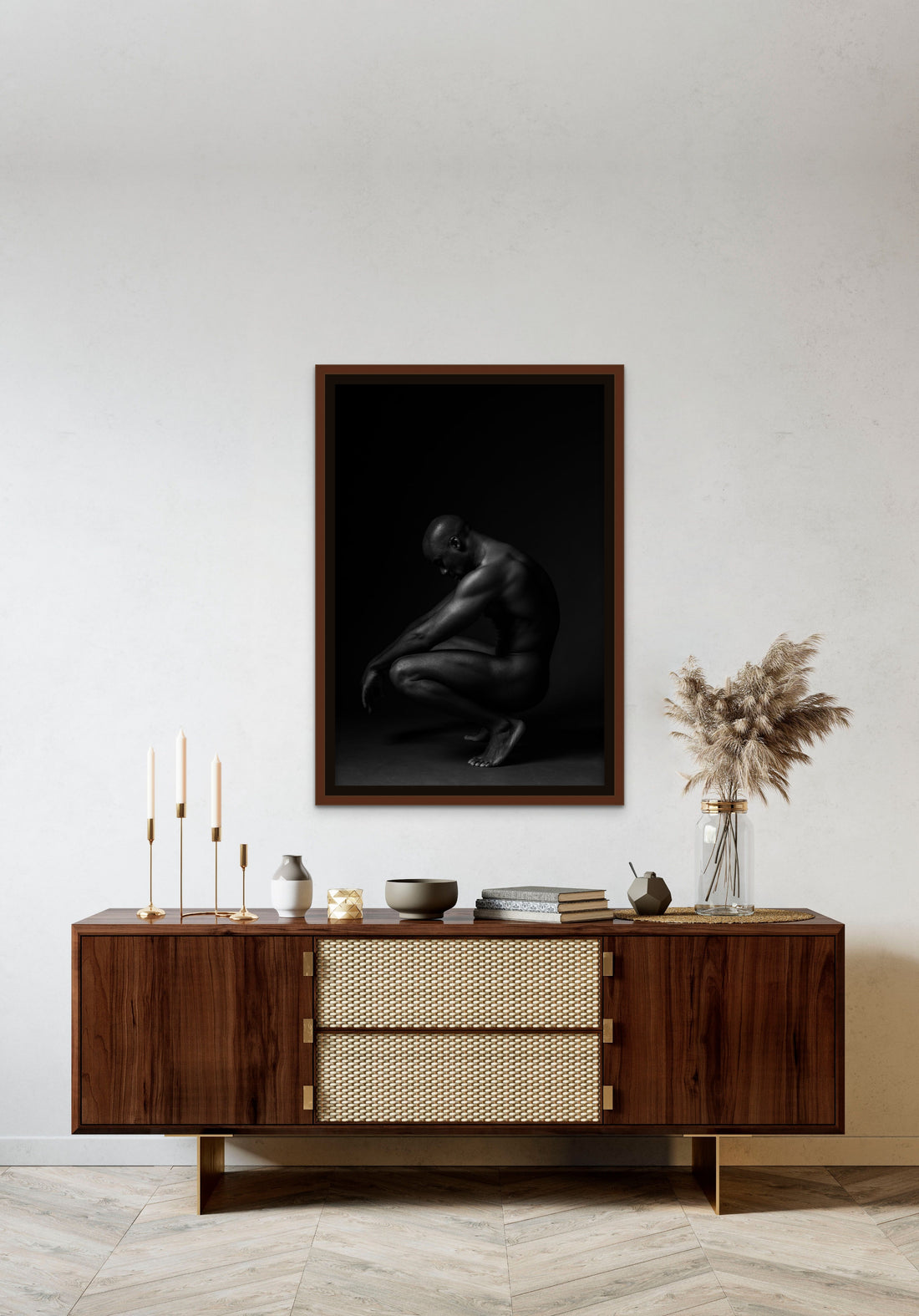 Framed canvas artwork hanging above a brown walnut credenza, the art is a black and white fine art photo showing a sideview of a nude black man crouching with arms resting on knees and head tilted down.