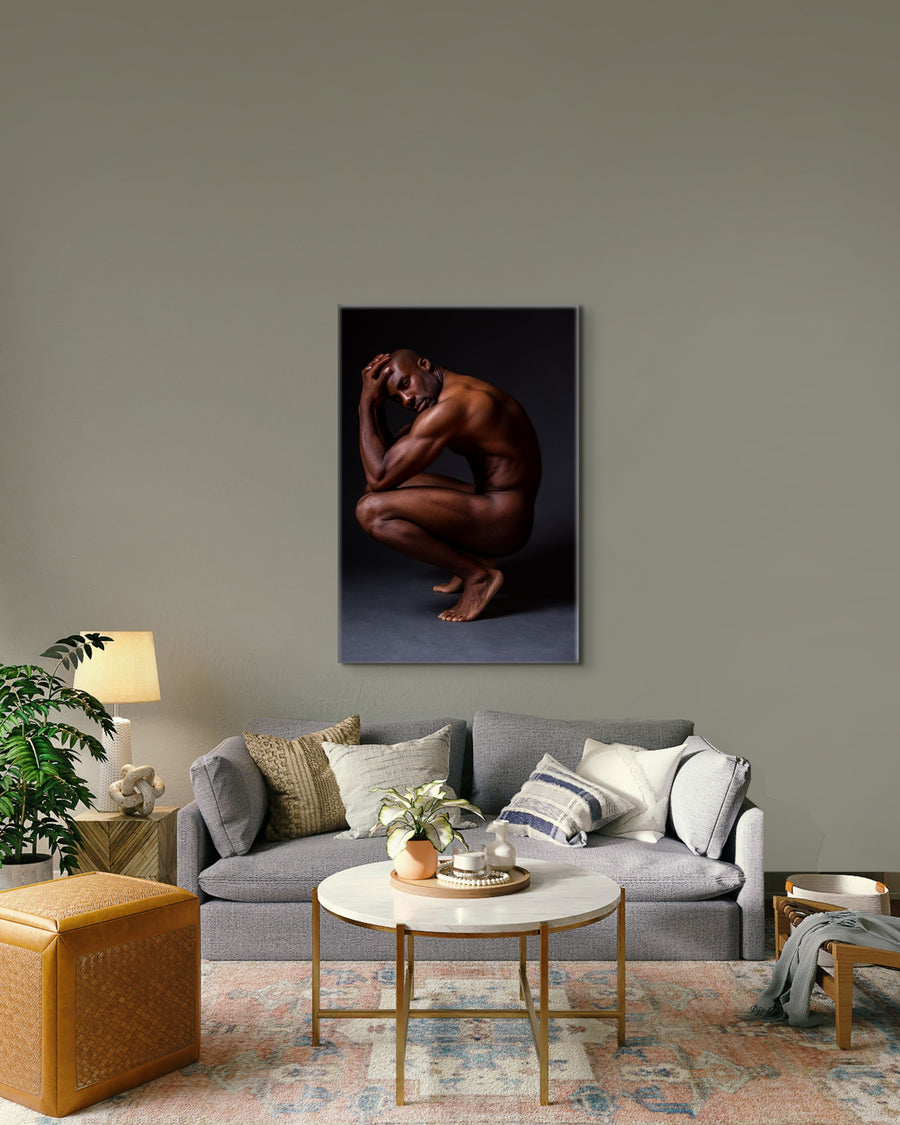 Photo on canvas artwork hanging above the sofa in a boho-glam style livingroom, the art is showing showing a sideview of a nude black man crouching with elbows on knees, hands on head, looking at the camera.