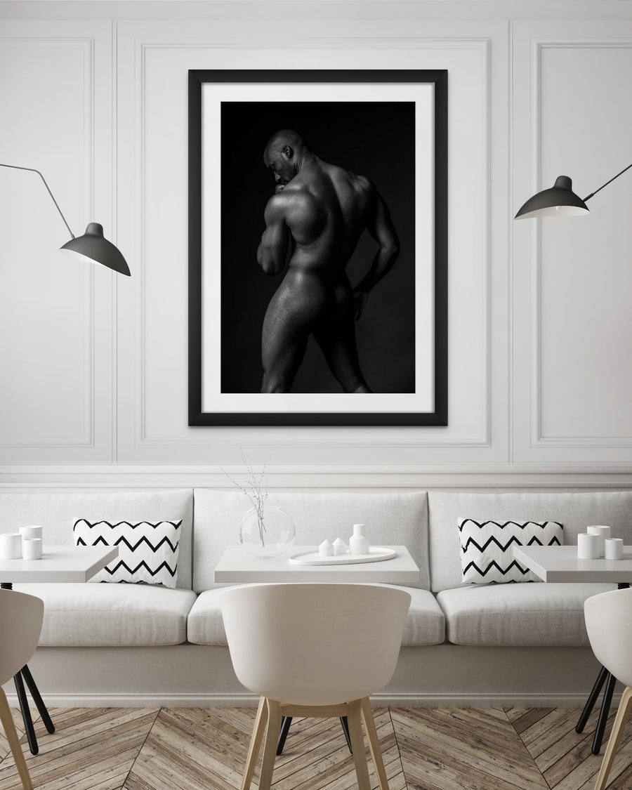 The inside wall of a modern classically styled restaurant with framed artwork hanging on the wall, the art is a black and white fine art photo of a nude black man posterior view from head to mid thigh with one arm bent and chin resting on closed fist.