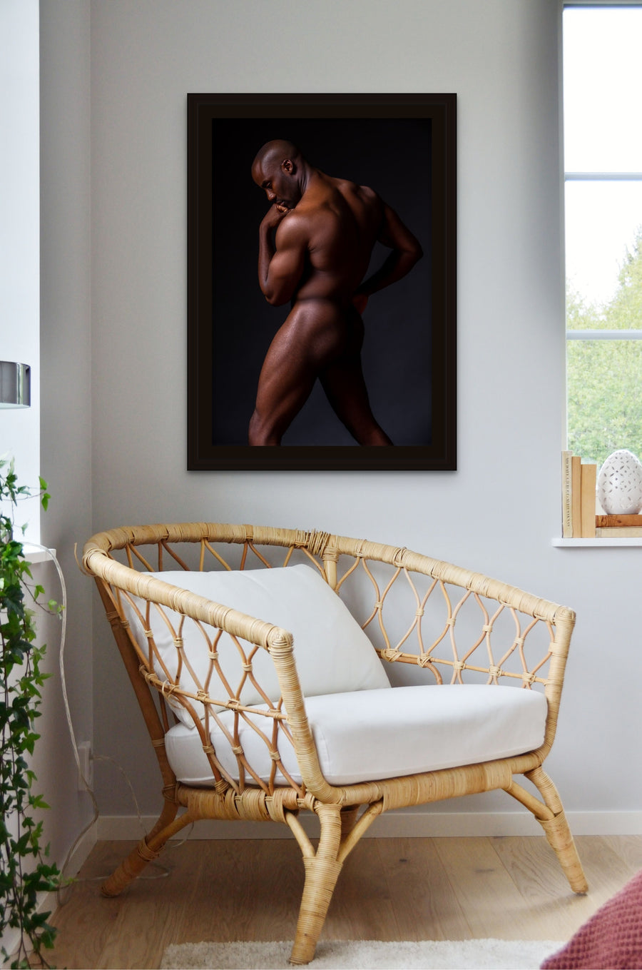 A rattan chair in a bright corner of a bedroom with artwork hanging on the wall above the chair, the art is a framed canvas photo of the sideview of a nude black man from head to mid thigh with one arm bent and chin resting on closed fist.