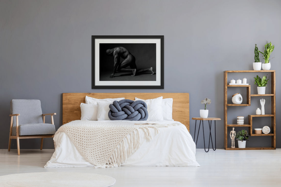 Framed artwork hanging above a light coloured wood headboard in a bedroom with natural and blue tones, the art is a black and white photograph of a sideview of a nude black man from head to mid thigh with one arm bent and chin resting on closed fist.