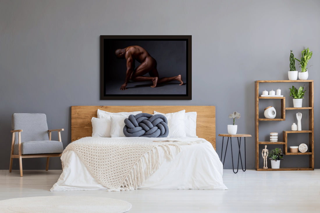 Framed canvas artwork hanging above a light coloured wood headboard in a bedroom with natural and blue tones, the art is a photograph of a sideview of a nude black man from head to mid thigh with one arm bent and chin resting on closed fist.