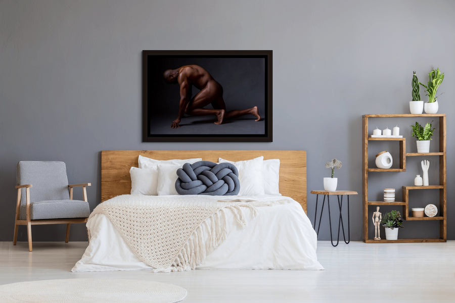 Framed canvas artwork hanging above a light coloured wood headboard in a bedroom with natural and blue tones, the art is a photograph of a sideview of a nude black man from head to mid thigh with one arm bent and chin resting on closed fist.