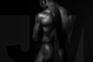 Black and white fine art photo of nude black man posterior view from head to mid thigh with arms crossed, looking downwards.