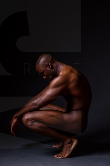 Fine art photo showing a sideview of a nude black man crouching with arms resting on knees and head tilted down.