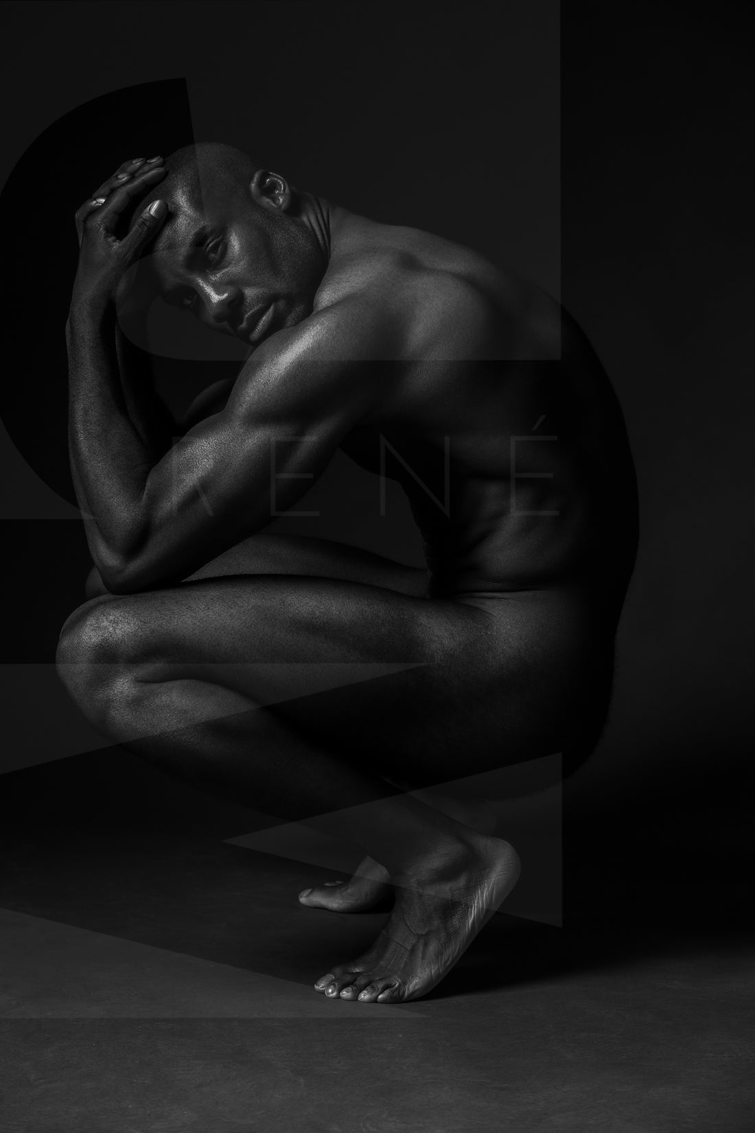 Black and white fine art photo showing a sideview of a nude black man crouching with elbows on knees, hands on head, looking at the camera.