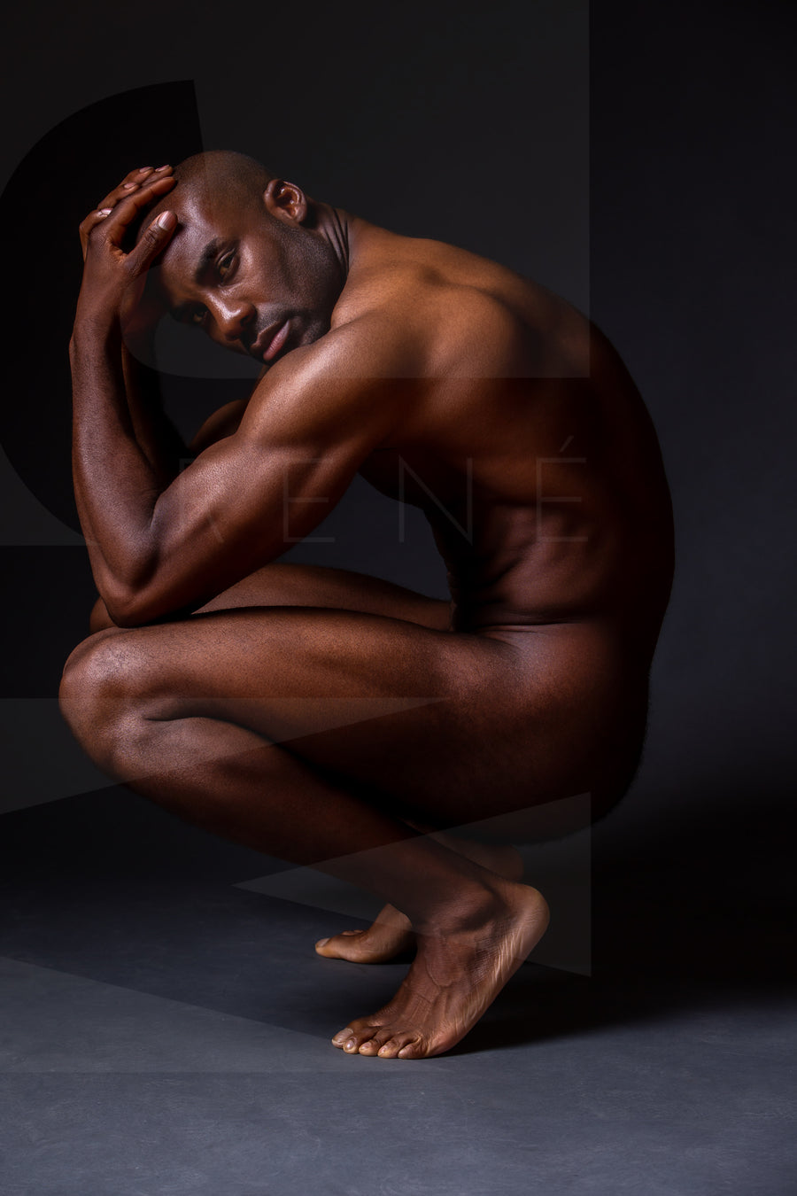 Fine art photo showing a sideview of a nude black man crouching with elbows on knees, hands on head, looking at the camera.