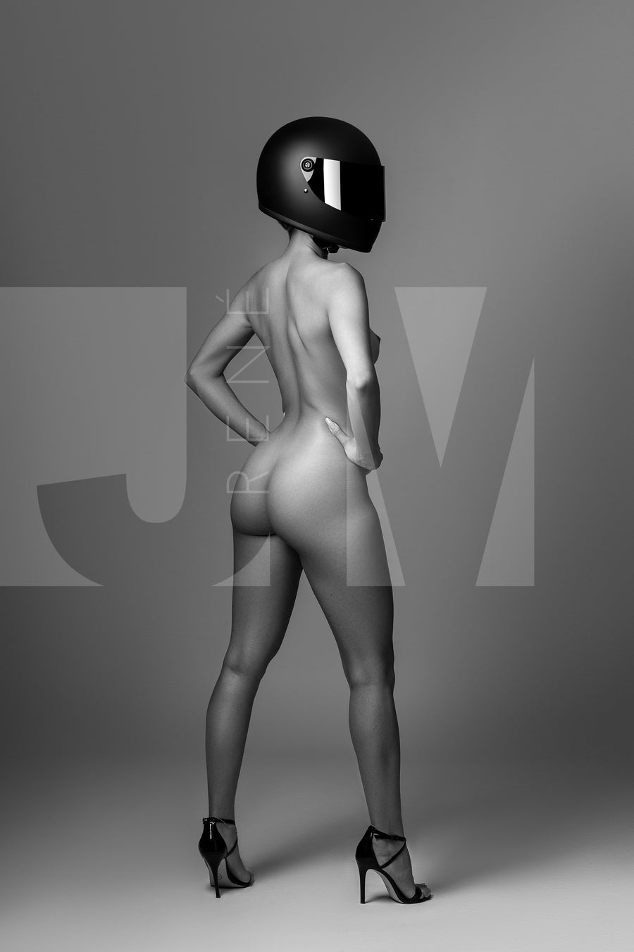 Black and white photo of confident nude woman wearing a black motorcyle helmet and black high heels standing with back to camera looking over shoulder.