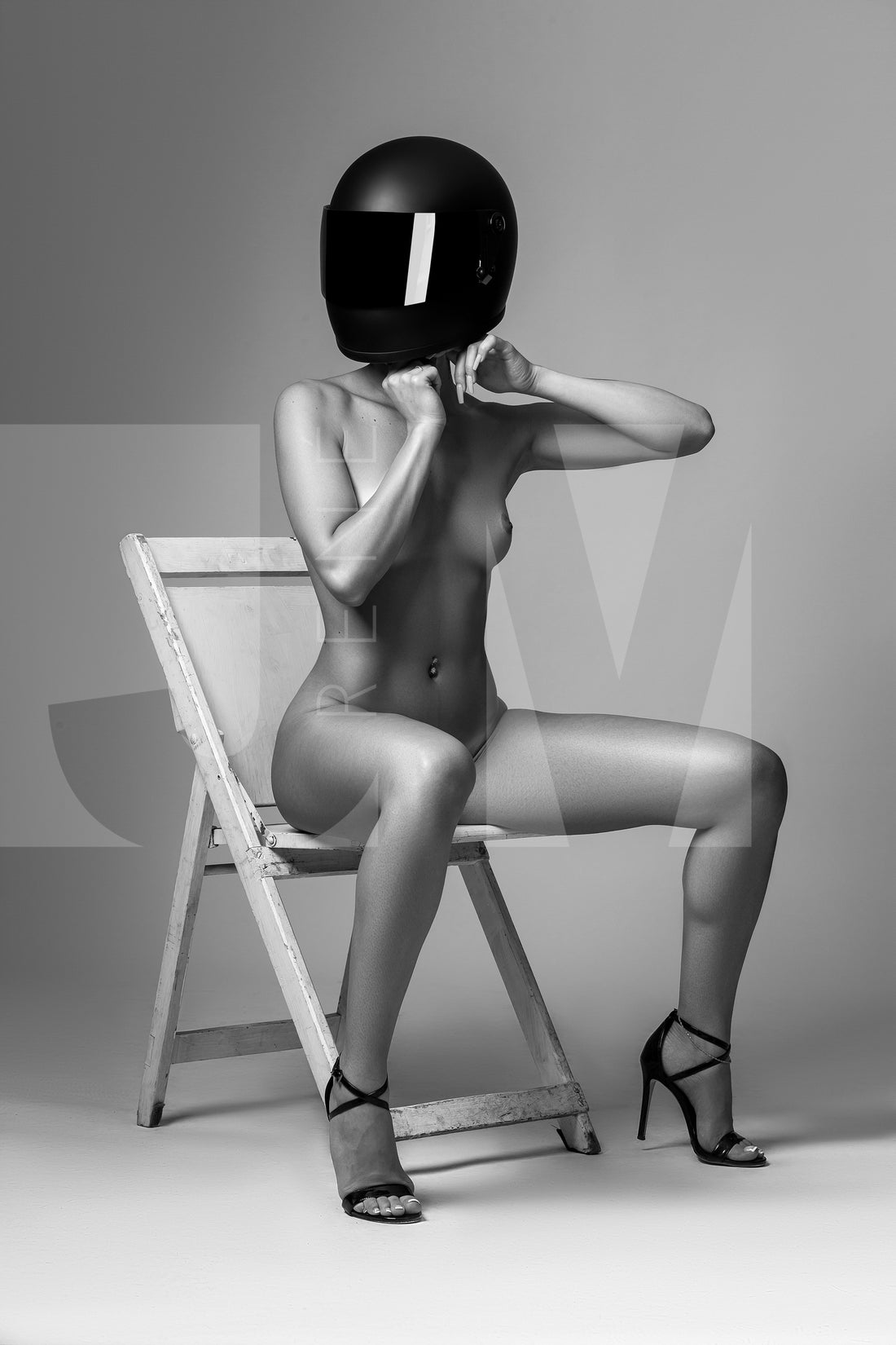 Black and white photo of nude woman wearing a black motorcyle helmet and black high heels sitting on chair adjusting helmet strap.