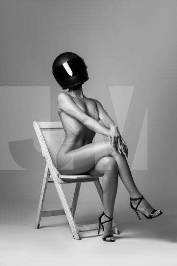 Black and white photo of nude woman wearing a black motorcyle helmet and black high heels sitting on chair with legs crossed, back arched and arms straight with hands resting on knee. 