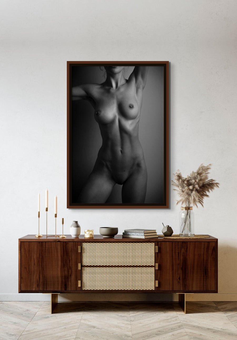 Framed canvas artwork hanging above a brown walnut credenza, the art is a black and white fine art photo of a nude woman standing facing the camera. The photo is cropped to show the model from her chin to her mid thigh. 