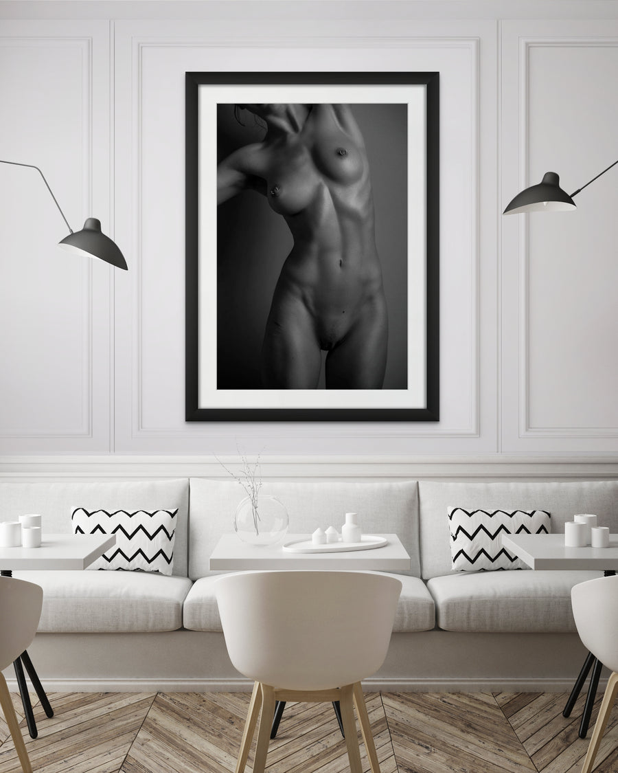 The inside wall of a modern classically styled restaurant with framed artwork hanging on the wall, the art is a black and white photo of a nude woman standing facing the camera. The photo is cropped to show the model from her chin to her mid thigh. 