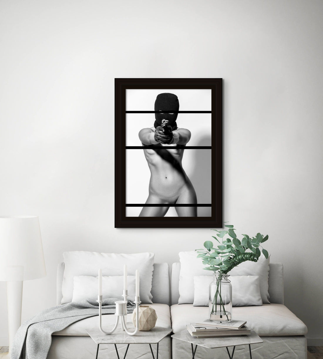 Framed fine art photo hanging above a sofa in a stylish livingroom of a nude woman wearing a black balaclava and black gloves holding a handgun, looking and pointing the gun directly at the camera.