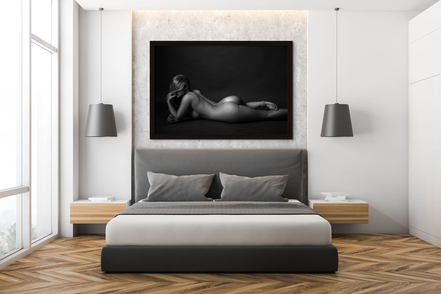 A clean and modern bedroom featuring a framed nude portrait of a woman lying on her stomach with her elbow bent and hand close to her face with is partially obstructed by her long hair. The model has a bent leg and has slightly rolled her hips to highlight the curve of her lower back, hips and buttocks. 