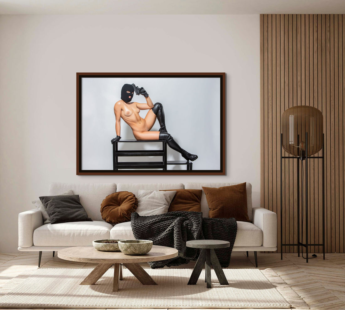 Framed fine art photo hanging above a sofa in a stylish livingroom of a nude woman wearing a black balaclava and black leather gloves and thigh high boots lounging on a bench with one arm bent and resting her handgun against her head in a seductive way.